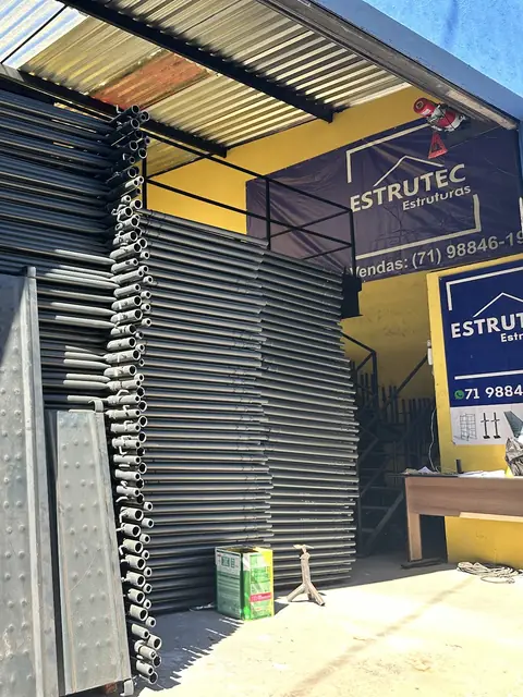 Painéis de andaime tubular empilhados na entrada da fábrica da ESTRUTEC em Salvador, com o banner de vendas ao fundo